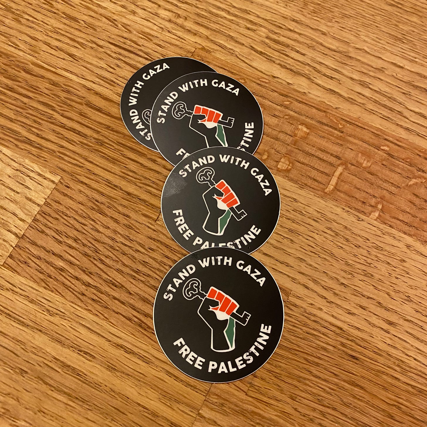 Four small black stickers on a wooden counter that say: Stand With Gaza. Free Palestine. In the center of each sticker is a fist holding a key in the colors of the Palestinian flag.