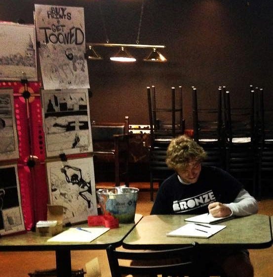 On the left, a display of black and white prints featuring fantasy characters and settings, next to an illustrated offer to draw people as cartoons. On the right. Dylan draws in a sketchbook.