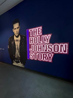 A large navy blue wall, with the neon-lit lettering of The Holly Johnson Story illuminated. Next to the words is a large, bigger than life-size black and white photo of Holly Johnson. He's wearing a black leather jacket and a white t shirt, and has dark hair.