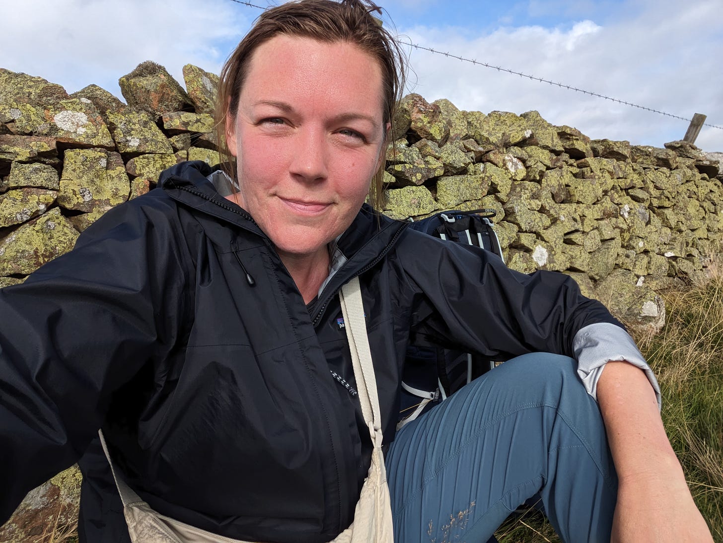 A red-faced sweaty Fiona sits by a dry stone wall with her rucksack next to her and waterproof coat on. She's on pilgrimage, pausing at the top of a big hill to rest.