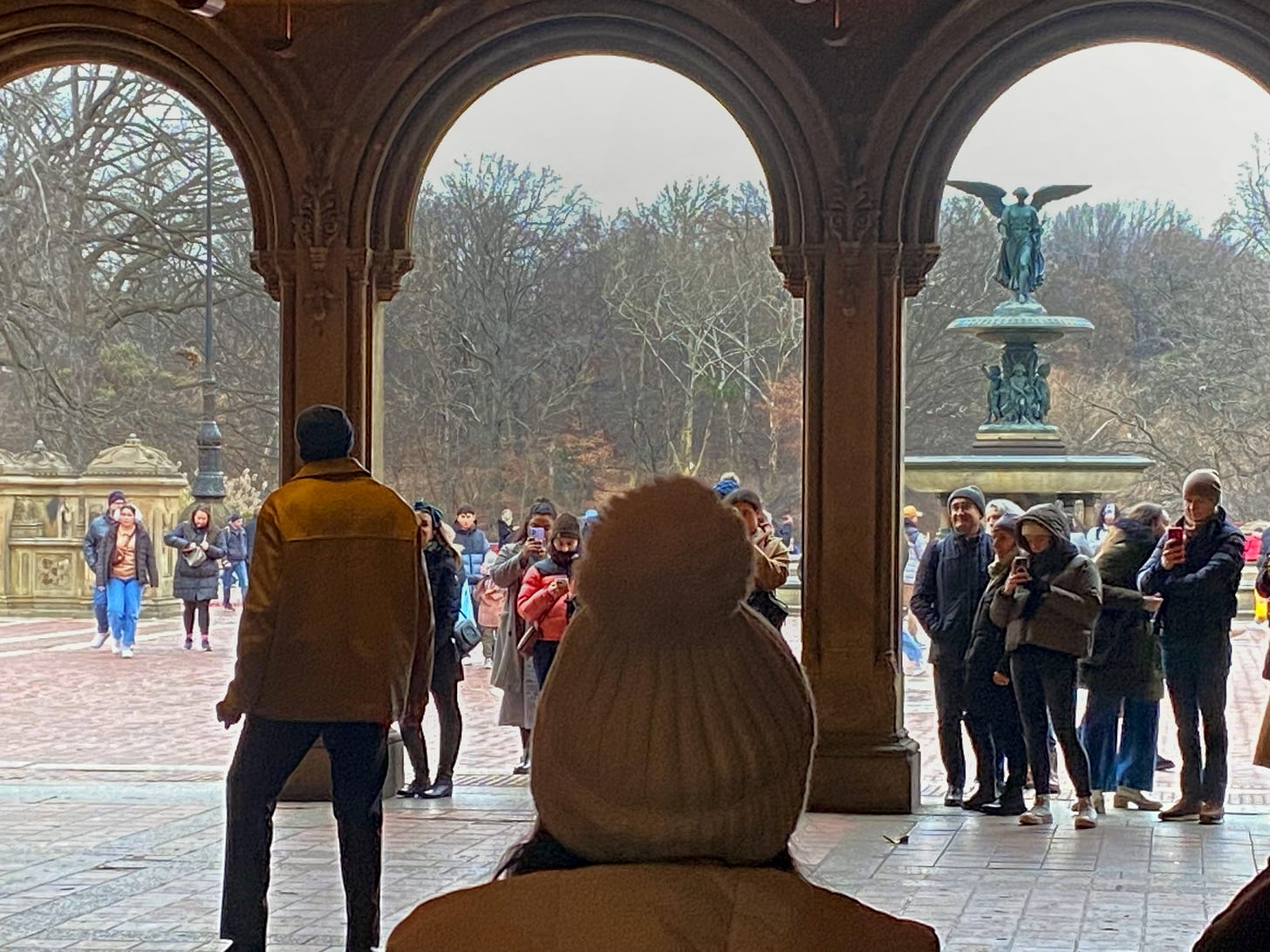 A man in a light-colored jacket sings jazz standards under the Bethesda Terrace. A crowd of bundled up New Yorkers stands watching and filming on their phones. The Angel of the Waters is visible through the right terrace arch.