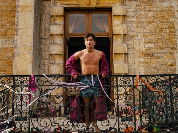 A young man wearing a purple robe, left open to reveal his bare chest, stands on a balcony covered in paper streamers.