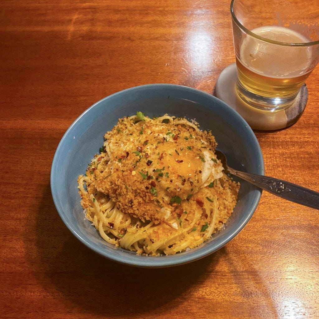A blue bowl of the pasta described above, with breadcrumbs, chili flakes and parsley over the top. The egg yolk is burst open and the fork is stuck beneath the noodles. On a coaster is a half-empty glass of IPA.