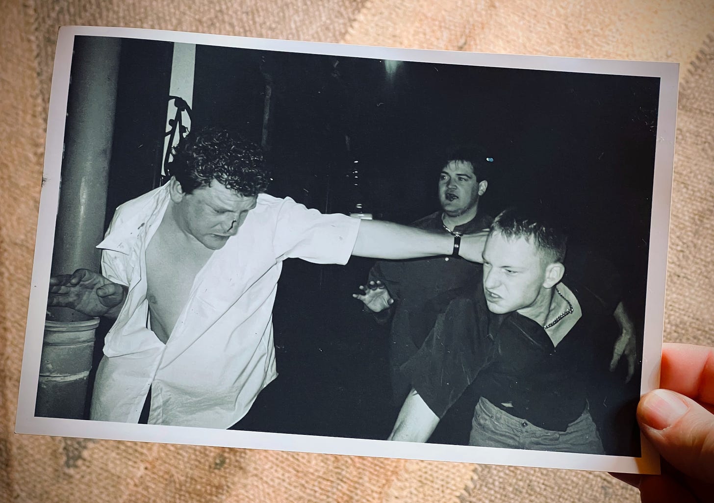 A hand holds up a photograph showing two young men mid brawl. One has just swung a punch and the other guy, bleeding from the nose looks dazed and in defence. In the background a guy with a swollen bleeding lip looks like he is half heartedly trying to stop the fight.