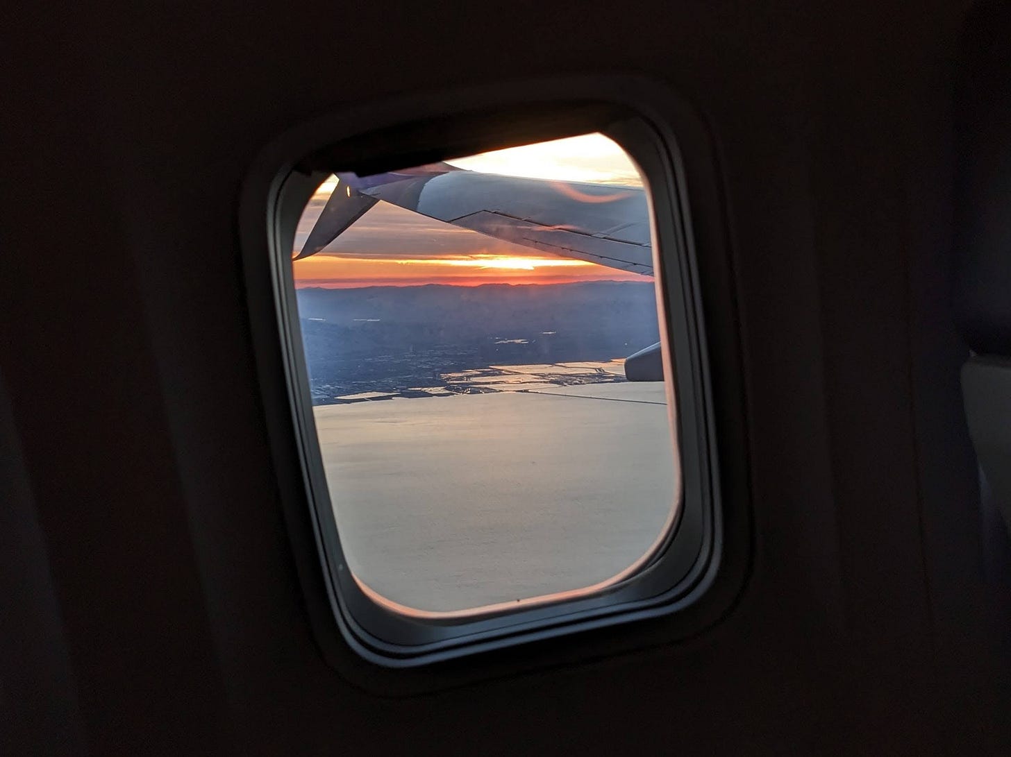 Photograph looking out of an airplane window at the sun rising over a city in front of water.