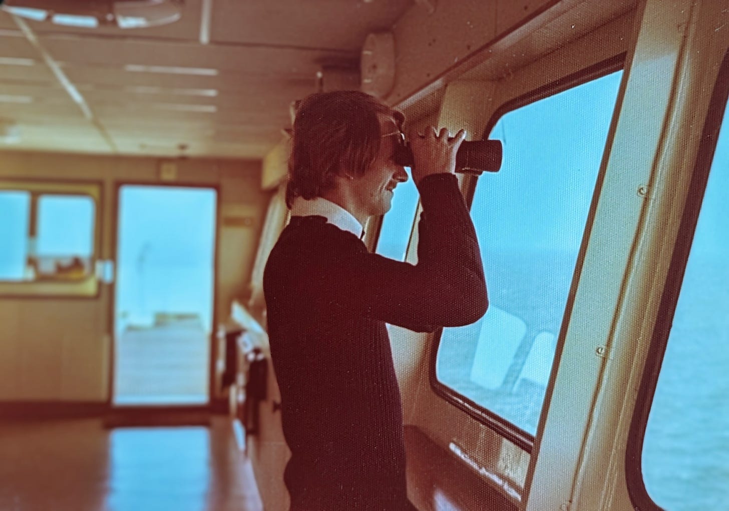 An old photograph of a young Merchant Navy Officer, dressed in a crisp white shirt and black jumper. He stares through large binoculars through the window on the ship's bridge, sunglasses propped up on top of them. he is smiling.