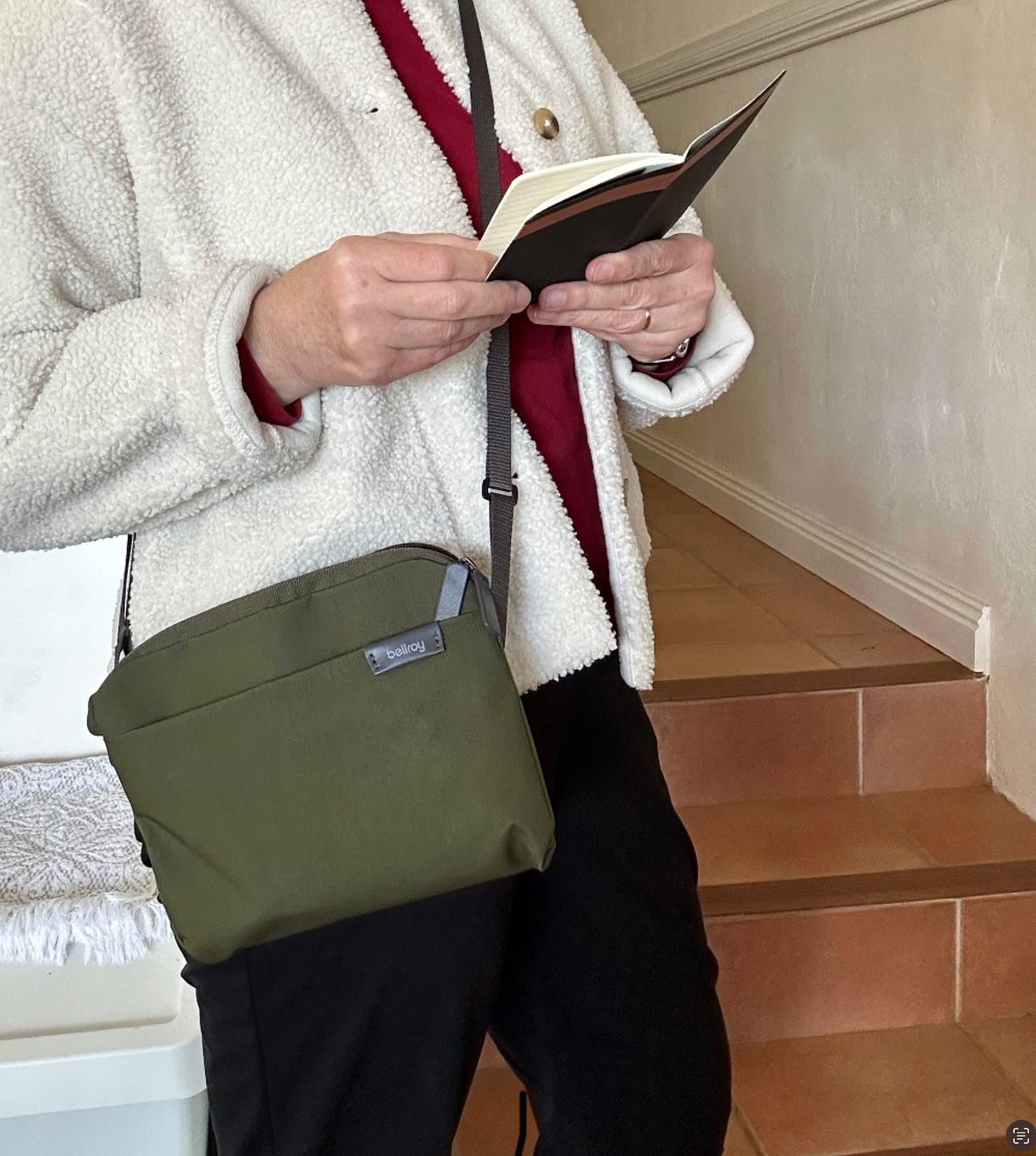 Torso of a person wearing black trousers and a fluffy cream jacket, standing in front of some stairs. They are wearing a khaki-coloured, compact cross body bag, and holding a opened, pocket sized black notebook.