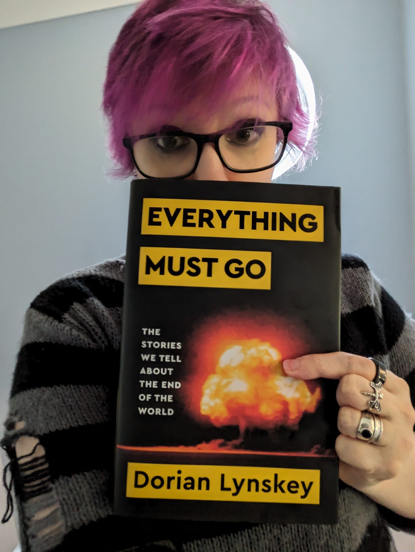 A woman with pink hair, a black and grey jumper, and rings, holding a copy of Everything Must Go by Dorian Lynskey, a large black volume with yellow-backed title and an image of an explosion