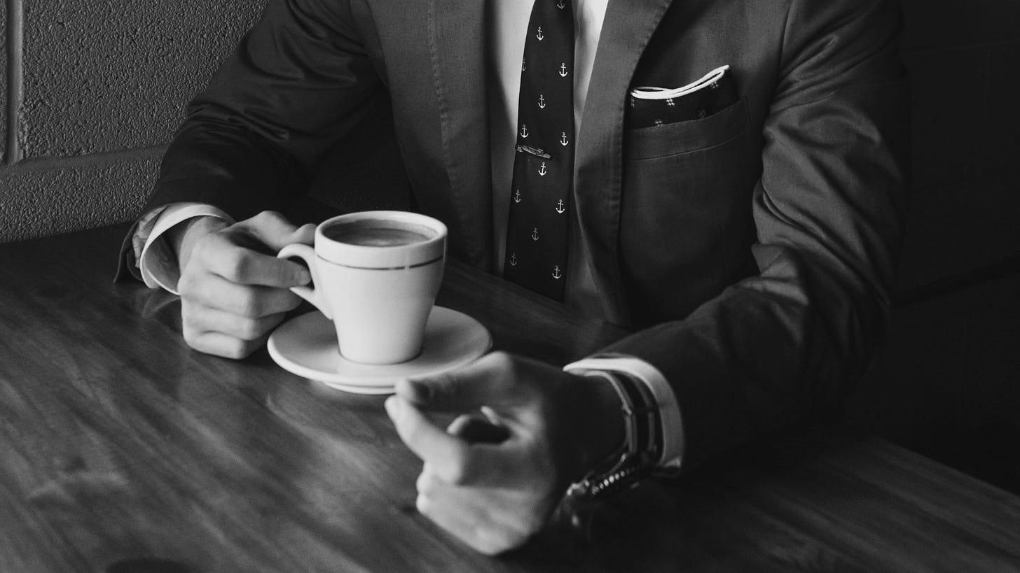 Close up of a person from the neck down wearing a suit sitting at a table with a cup of coffee in front of them. Their right hand holds the cup while their left reaches towards the camera