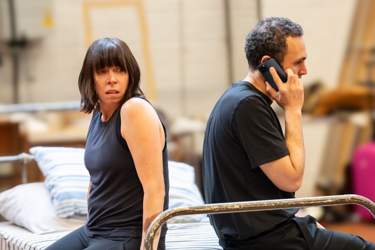 Two actors sat at the end of a bed, facing in opposite directions. The woman has brown hair and an upset expression. The man has short curly hair and is holding a phone to his right ear.