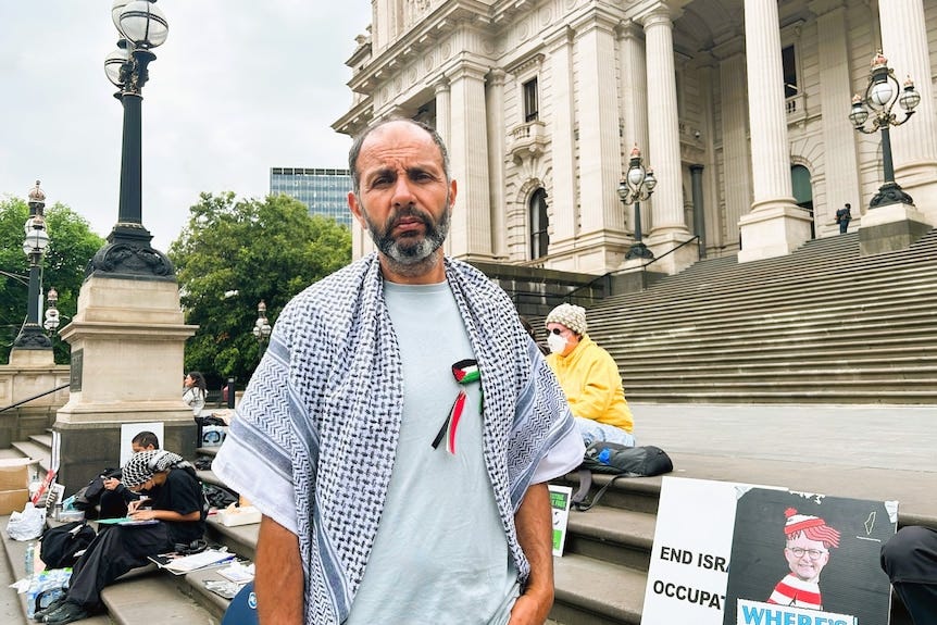 A man stands wearing a palestinian scarf, therer are signs behind him and parliament house is shown in the background.