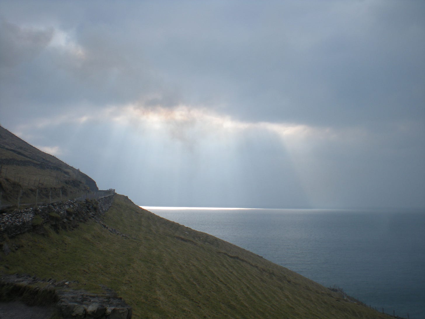 A narrow road runs around the edge of a green hill, with rays of light shining down on a distant portion of the sea. 