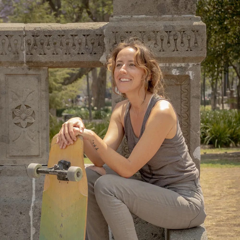 A smiling woman crouches next to her skateboard.