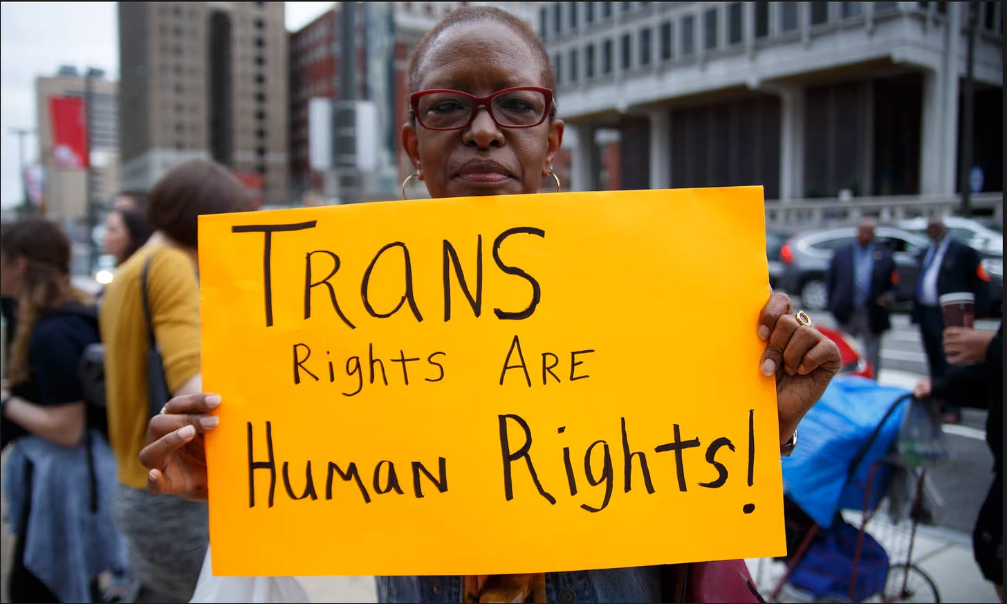 An elderly black woman holds a yellow sign that says "Trans Rights Are Human Rights" in big lettering. She is standing outside of a city hall at a protest