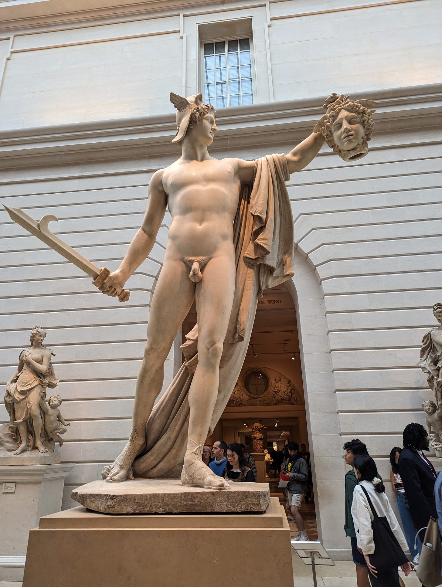 the Greek hero Perseus poses with the head of the Gorgon Medusa. In one hand, he holds a weapon. In the other, he holds Medusa's head. He's depicted as mostly nude with a robe or cape draped over his arm.