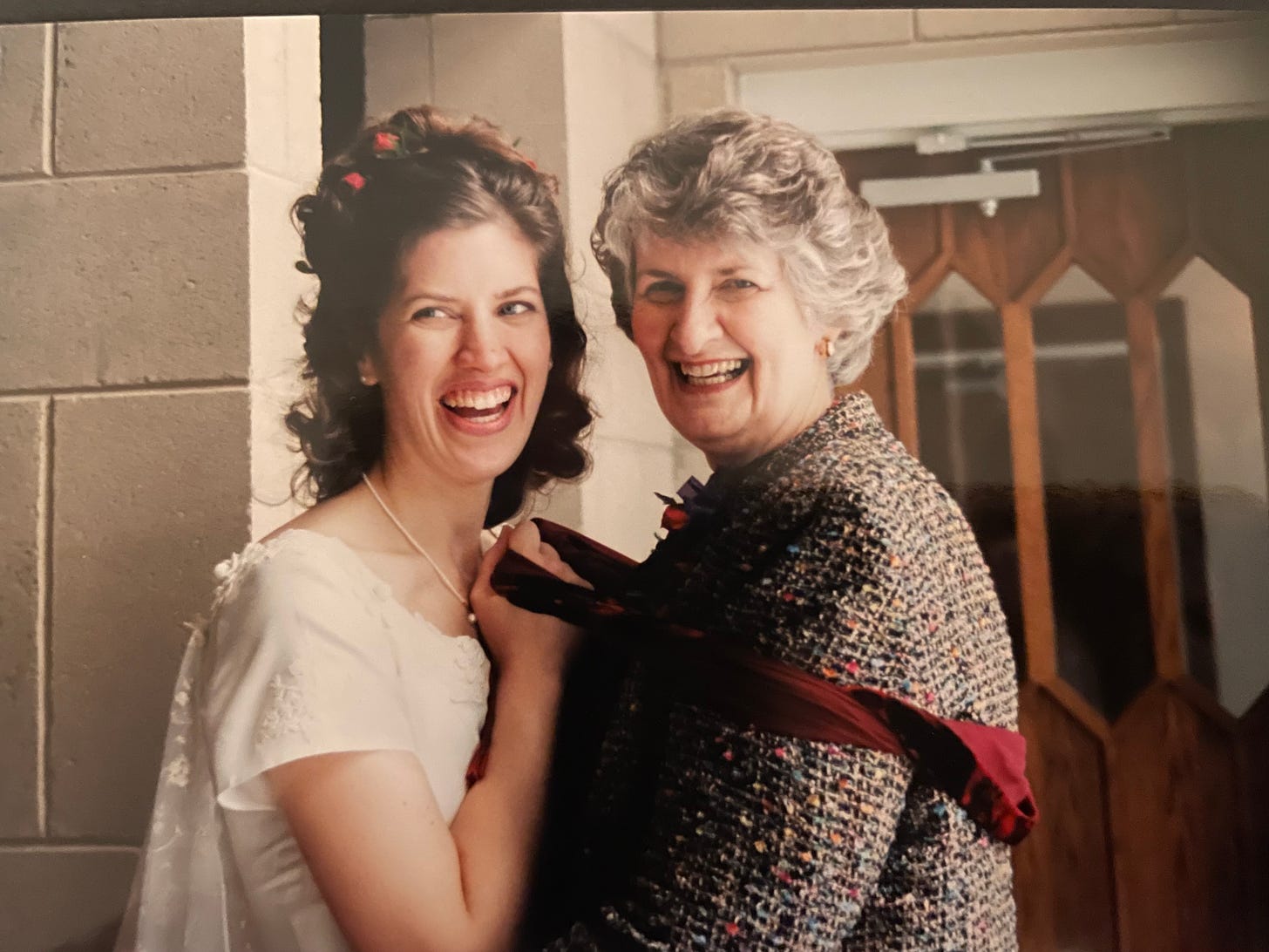 A young-ish woman (me) in a wedding gown uses a shawl to pull close a middle-aged woman (Mom) in a blazer