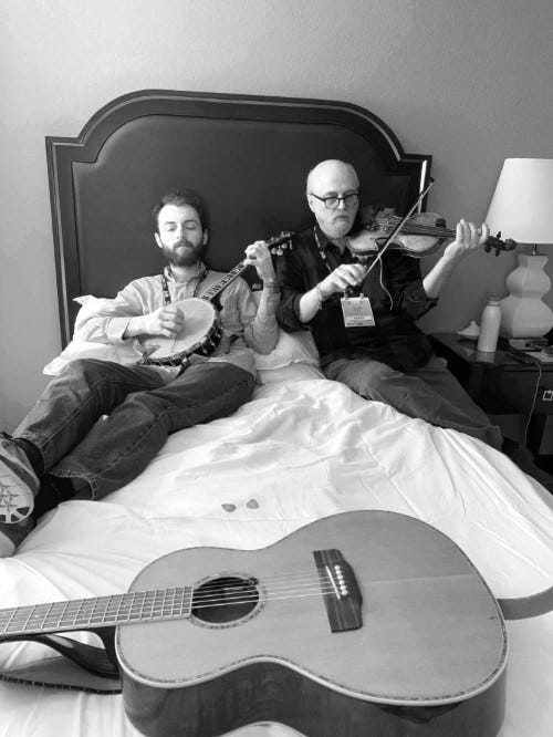 Two men playing fiddle and banjo, practicing while sitting on a hotel bed.