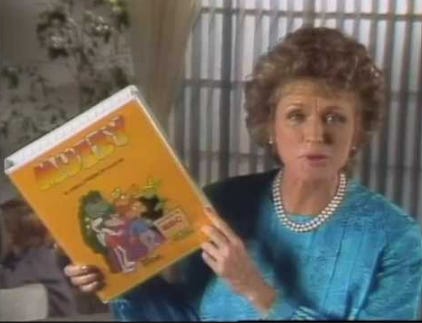 A screencap of the Muzzy commericals. A woman in a very blue, pleated dress and pearls is holding a huge VHS tape box of Muzzy language-learning tapes. Seul les enfants des années 90 sauront.
