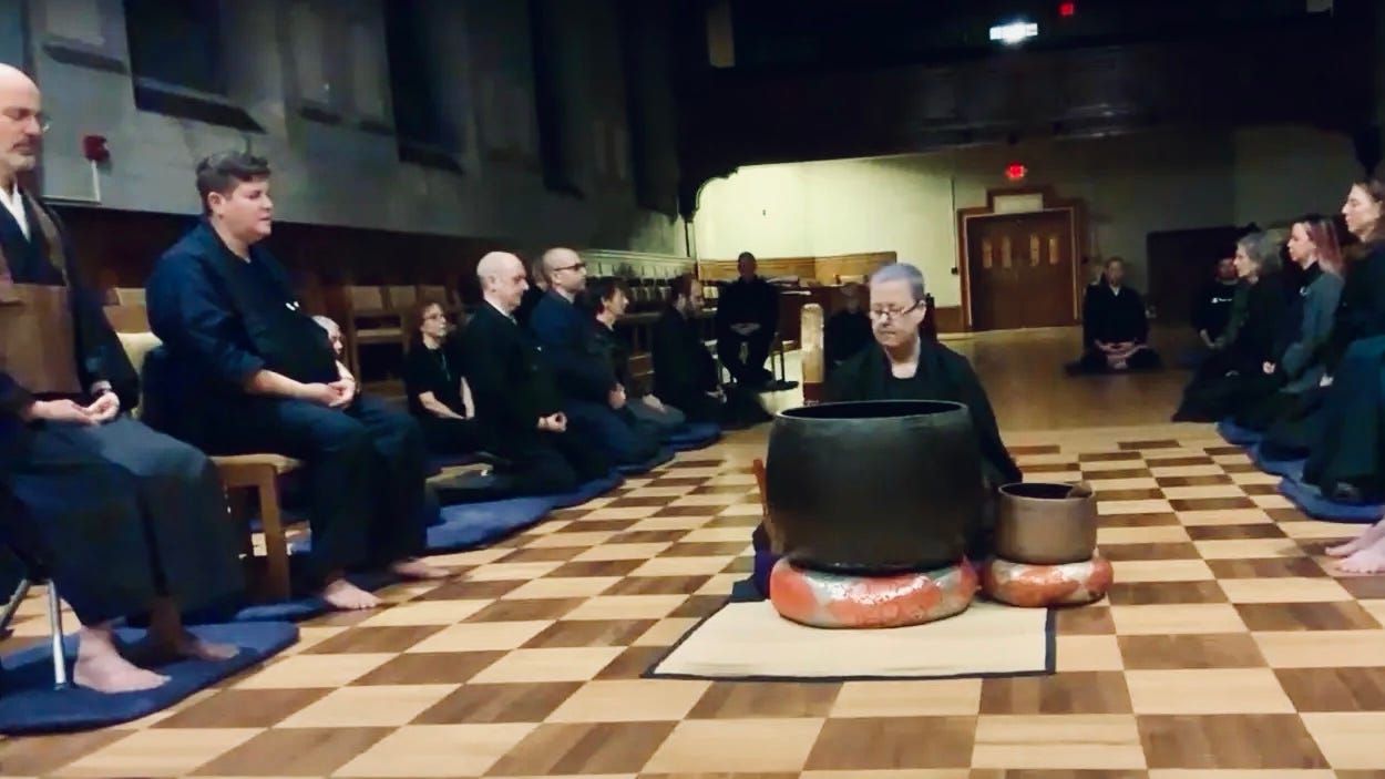 Gessho, a white woman in zen robes with blue hair, sits behind a large keisu bell, holding up a striker. On both sides of her, rows of Zen teachers and students sit in meditation.