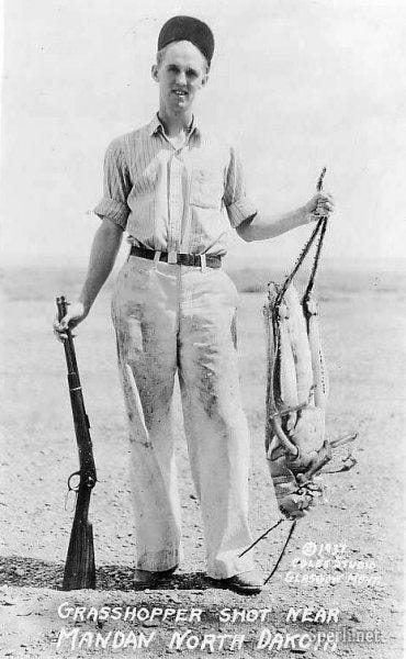 A black and white photo of a man standing in the field with a rifle, holding what appears to be an enormous dead grasshopper