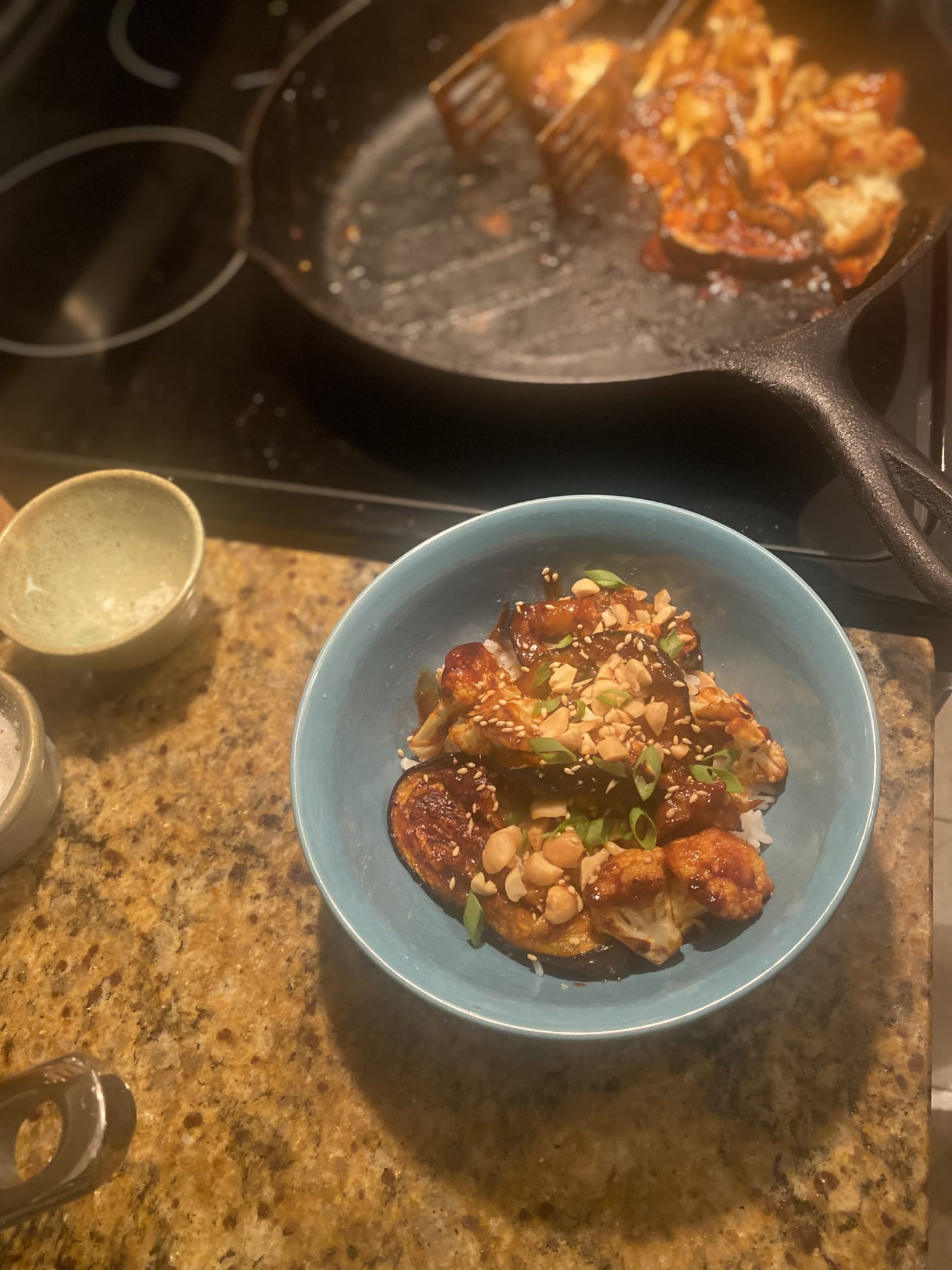 A blue bowl on the counter full of rice and eggplant and caulilower in a deep red glaze. On top are chopped peanuts, sesame seeds, and chopped green onions. In the background on the stove is a cast iron pan with the remainder of the gochujang-glazed eggplant & cauliflower.