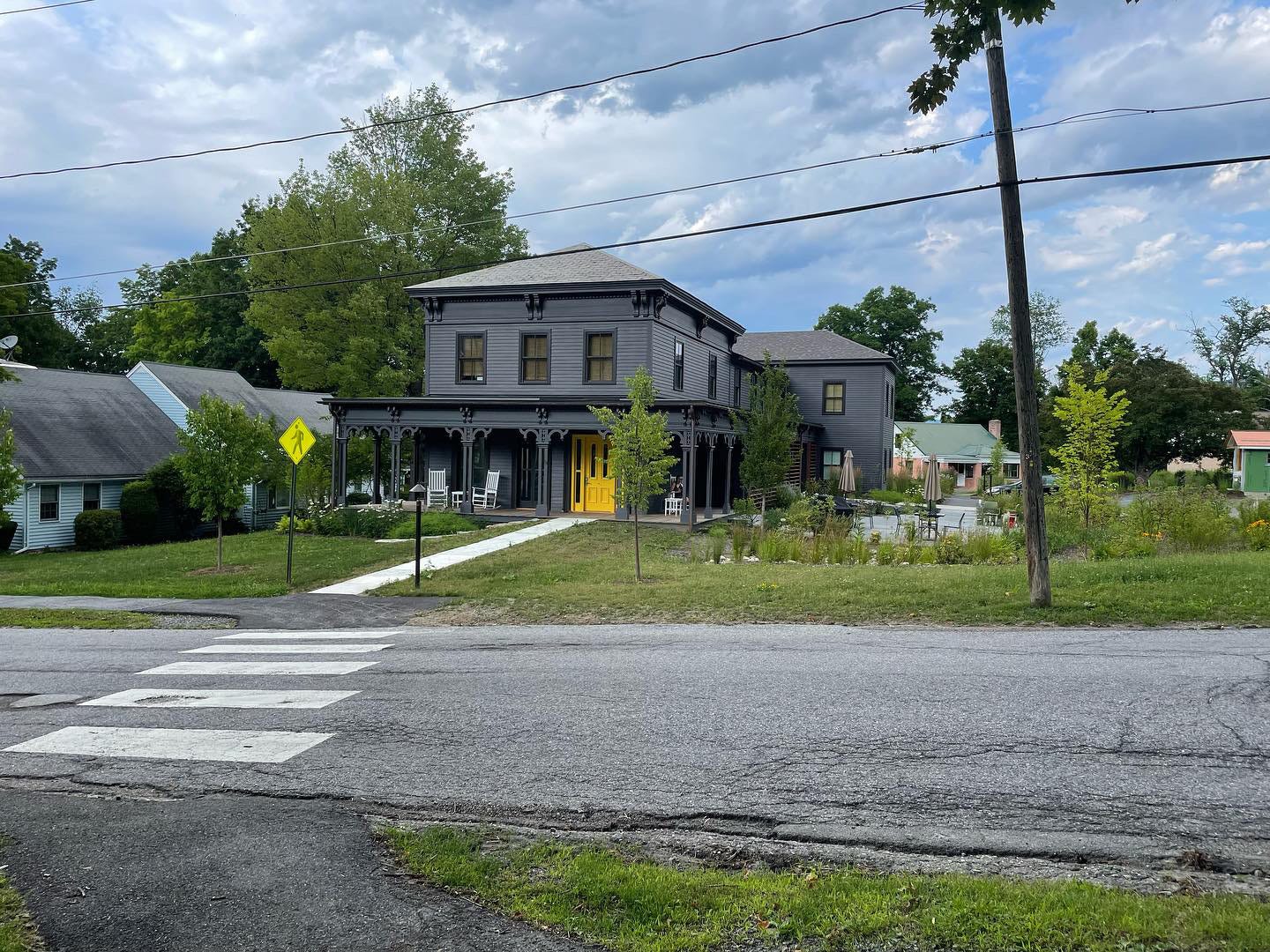 A gray farmhouse with a porch and sidewalk in front showing a crosswalk and front yard and a blue sky filled with clouds. 