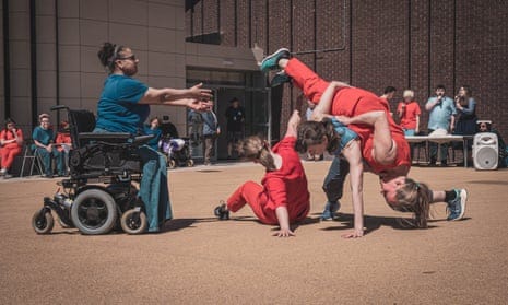 A woman of color in a powerchair reaches towards three white dancers entangled mid-dance.