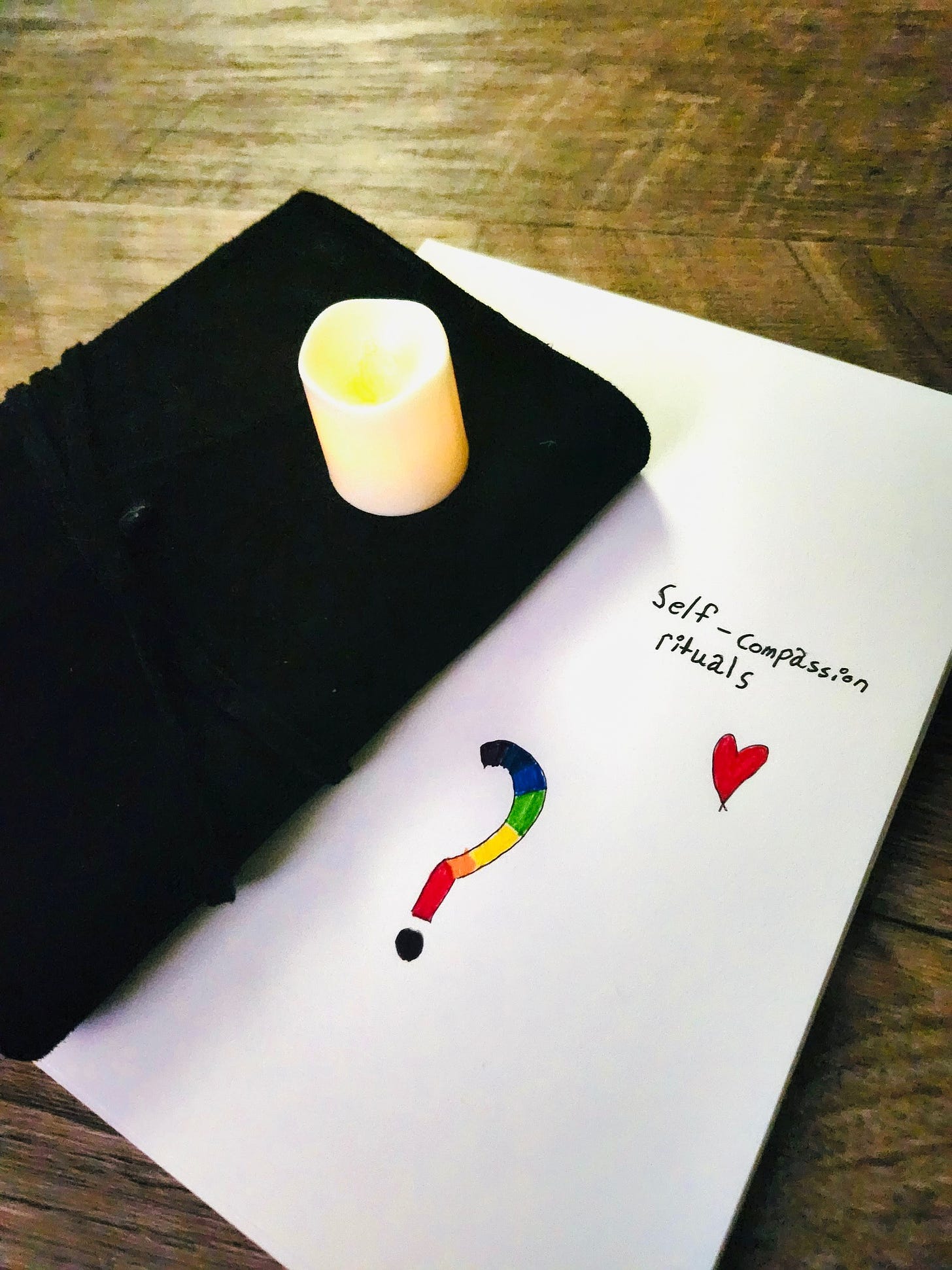 "Art journal page that says 'Self-compassion rituals,' featuring doodles of a rainbow question mark and a heart. Sitting on top of the art journal are a white LED candle and a black leather journal.