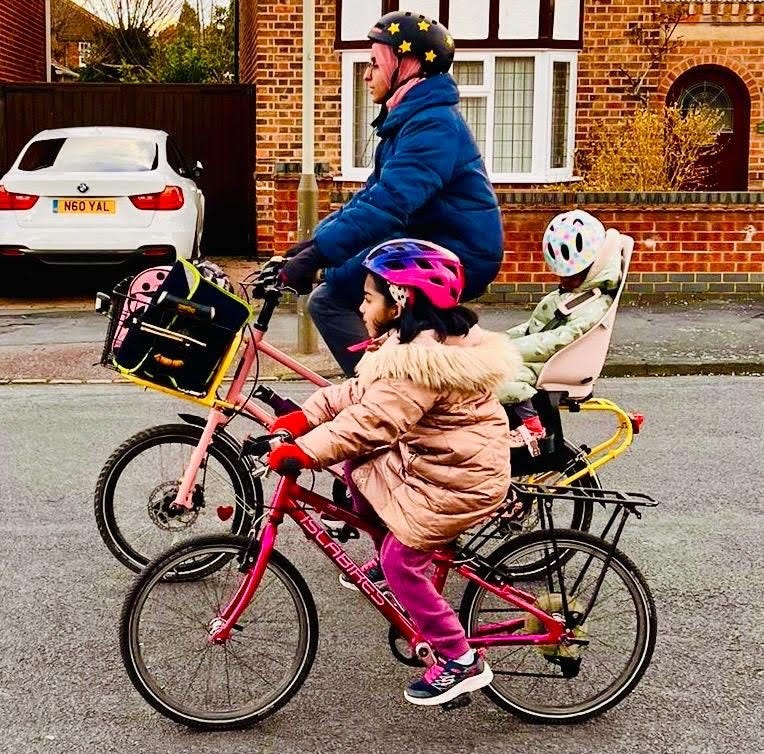 Cycling in Ramadan - Sarah, a Muslim woman wearing a Hijab under her bike helmet, cycling a cargo bike with a toddler in a rear child seat, next to her older daughter who pedals her own Islabike