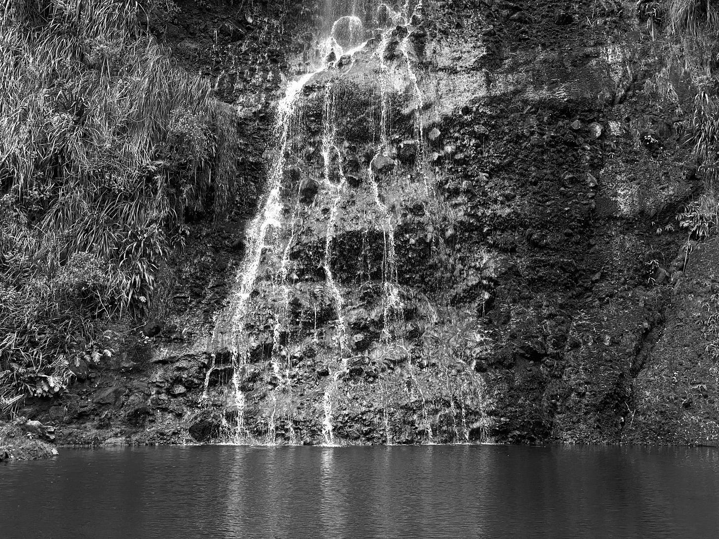 Photo of a small waterfall branching and flowing in several thin strands down a vertical, rocky hillside into a calm pool.