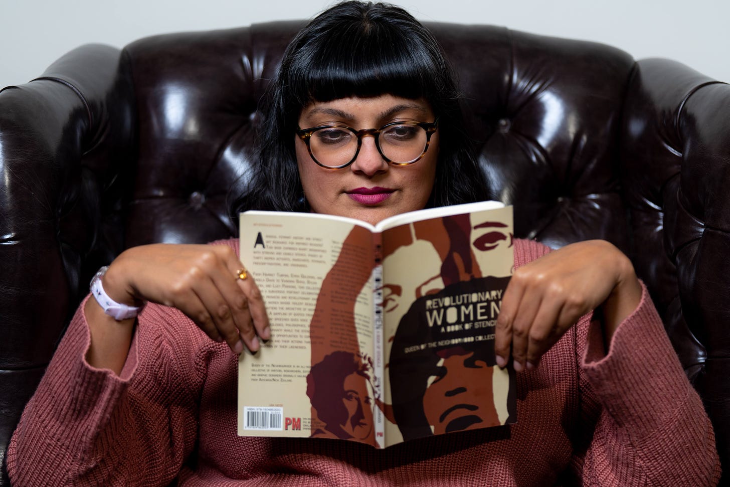 Nisha in a brown leather chair reading "Revolutionary Women: A Book of Stencils". She wears glasses and a pink sweater.