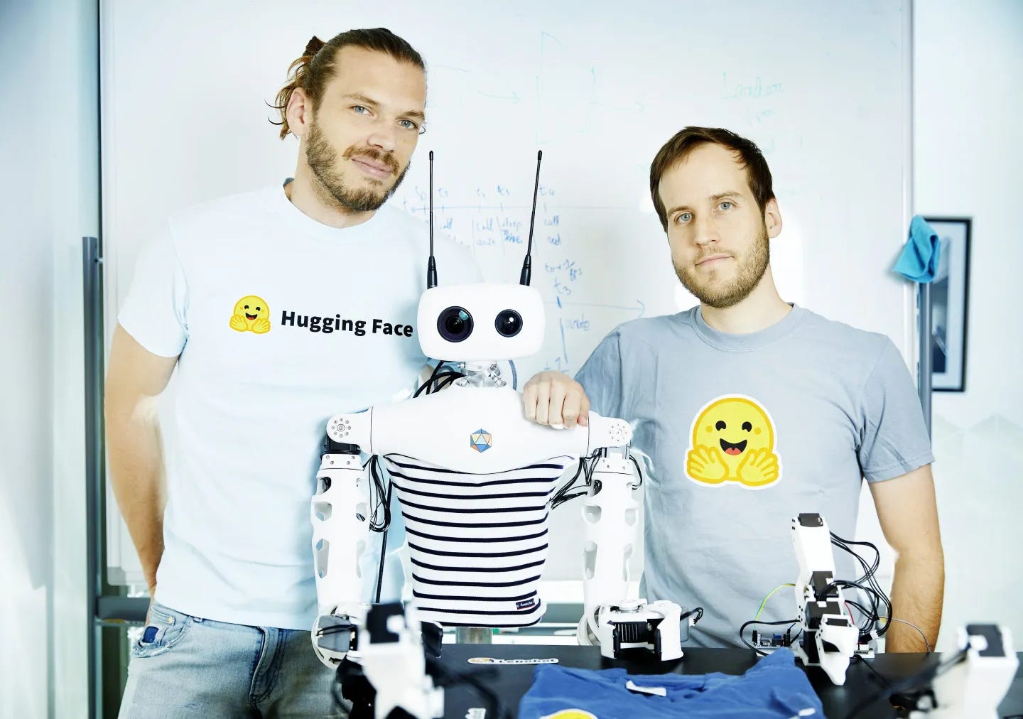 Simon Alibert and Rémi Cadene, robotics researchers at Hugging Face, pose wearing T-shirts with the Hugging Face logo as they stand alongside Reachy 2, a humanoid robot built by Pollen Robotics.