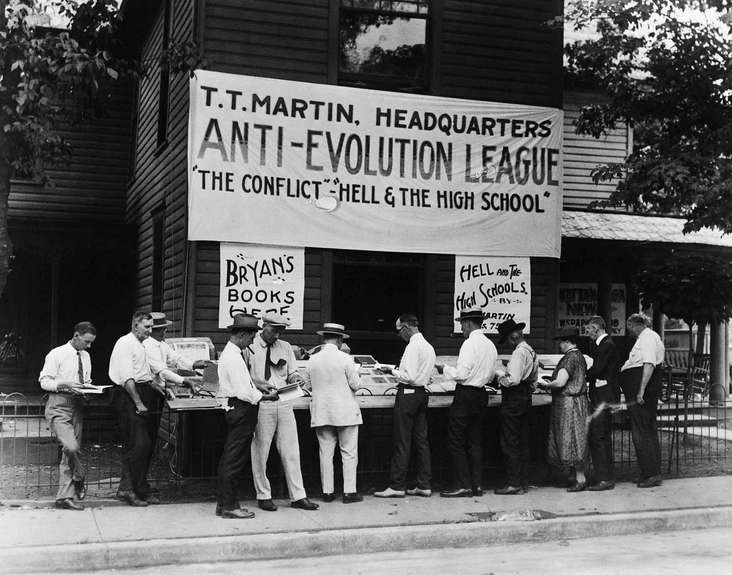 Black and white historic photo of people at a book display outdoors with a large banner reading "Anti-Evolution League: The Conflict Between Hell and the High School"