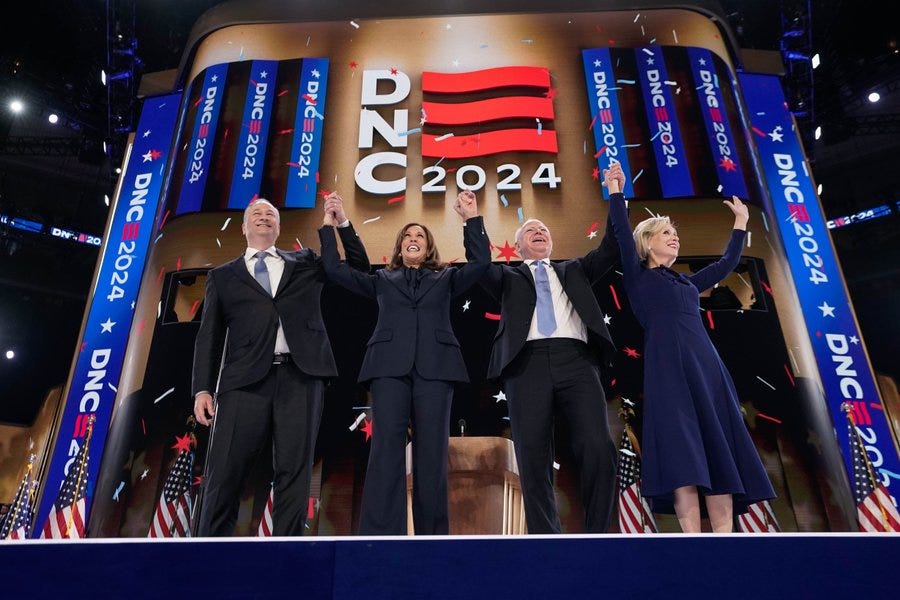 Photo of Democratic Presidential Nominee Vice President Kamala Harris, Second Gentleman Doug Emhoff, Minnesota Governor Tim Walz and First Lady Gwen Walz holding hands in the air as confetti falls to the ground on the final night of the 2024 DNC. The Democratic National Convention 2024 logo is displayed on a large screen in the background.  