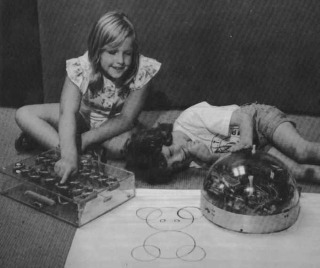 A photo of two smiling children playing with a small domed machine on the ground, with the child on the right tapping a control panel and the child on the left laying on the ground next to the robot.