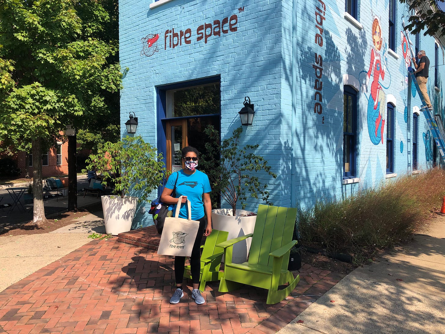 Kristen stands in her Carolina Panthers colored home period t-shirt in front of the doors of Old Town Alexandria yarn shop Fibre Space in August of 2020. Because it’s August of 2020, she’s still masked outdoors