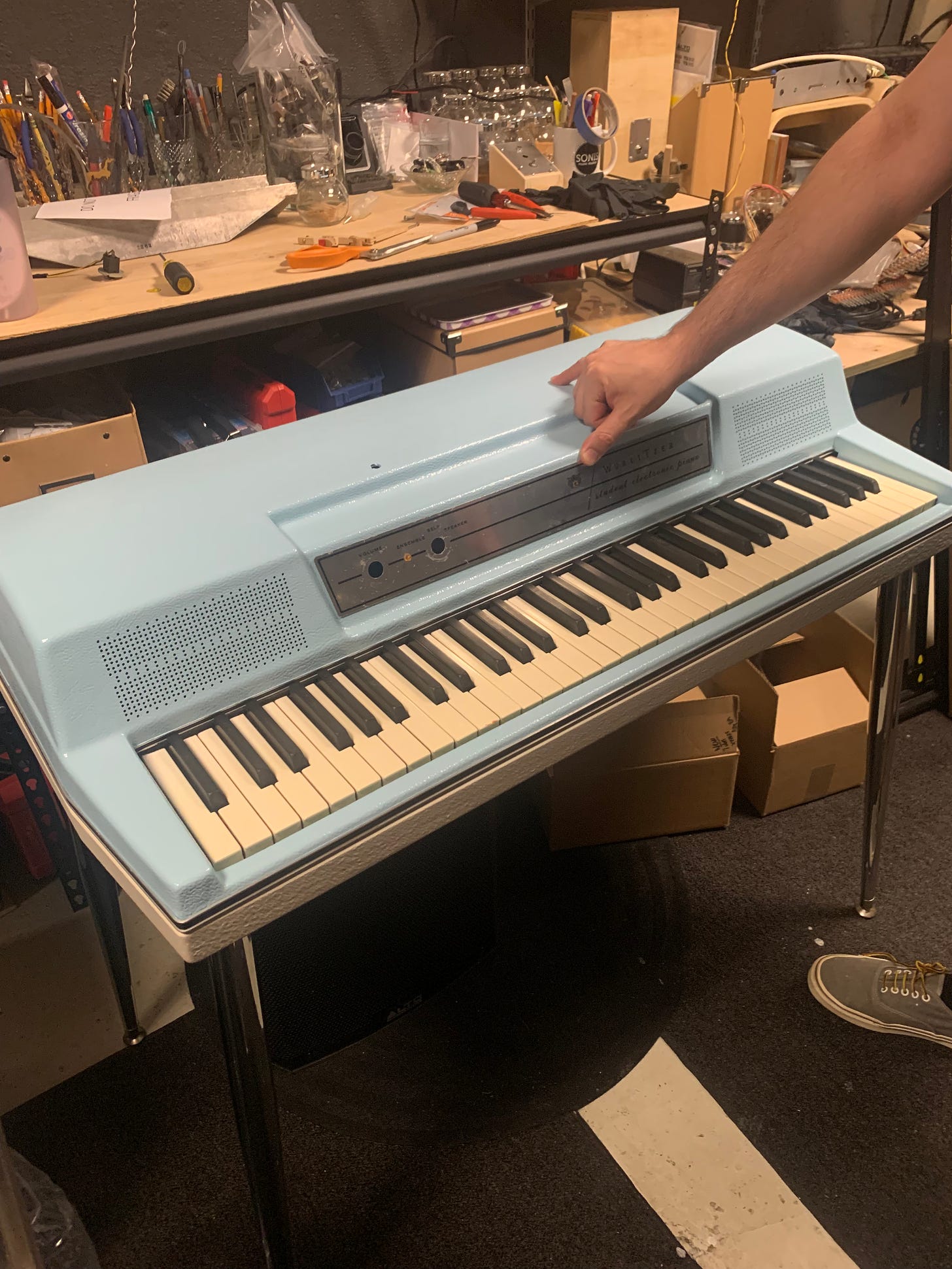 The blue Wurlitzer in the shop, in front of a shelf that has tools and a harp on it. Jon is holding the faceplate on with his thumb.