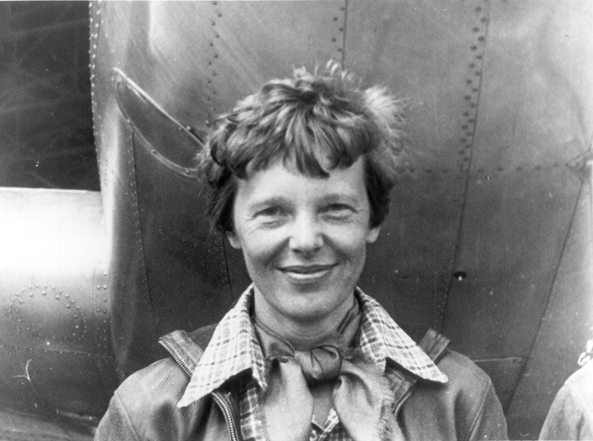 Headshot of Amelia Earhart. She is standing under the nose of her plane, although most of the plane is not in the picture.