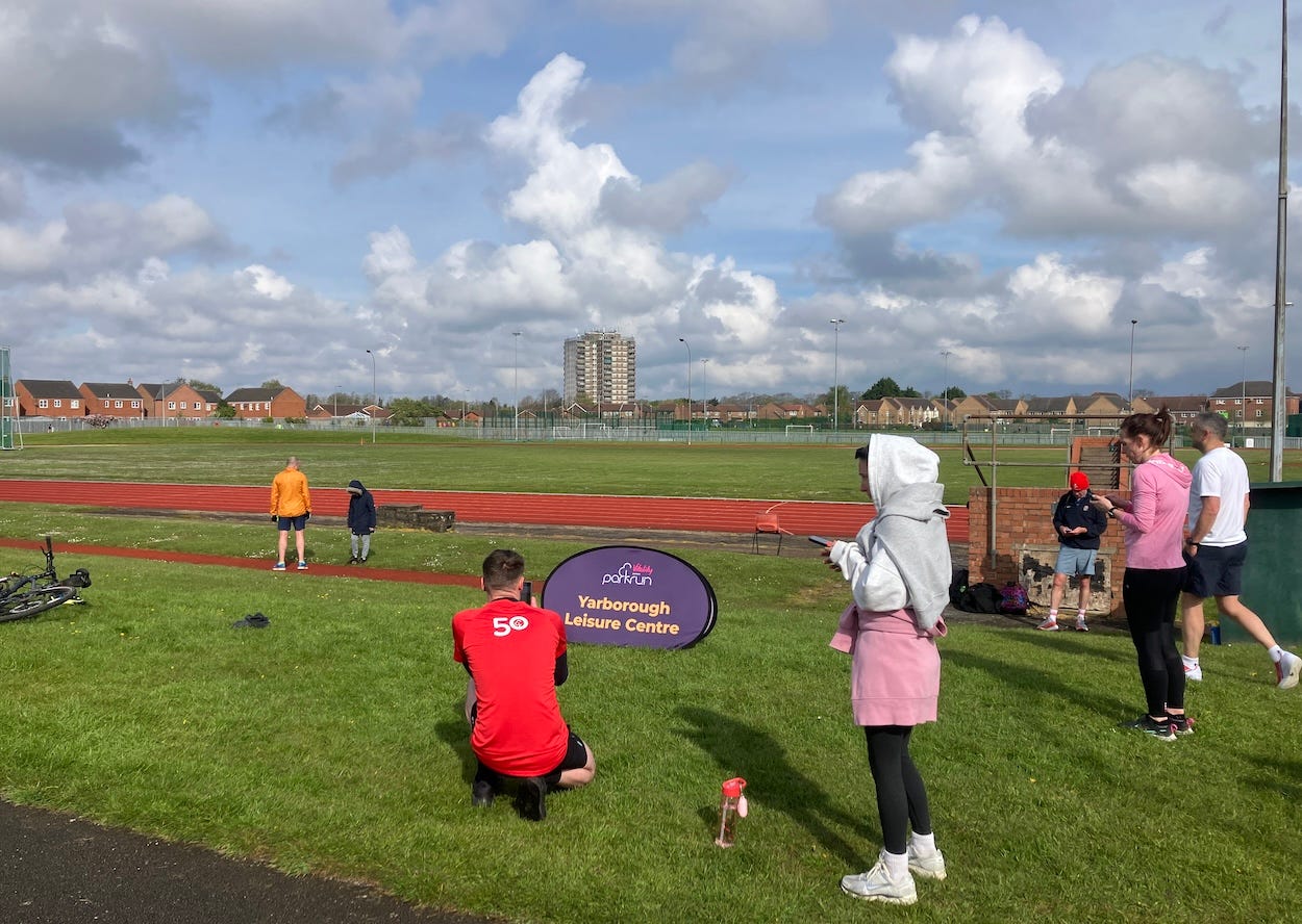 A group of people congregates near a sign at an outdoor athletic track with red surfaces within a suburban park. Partly cloudy skies hint at recent weather changes, and a mix of residential structures can be seen in the background.