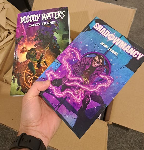 Franks' hand holding copies of Bloody Waters and Shadowmancy over an open box.