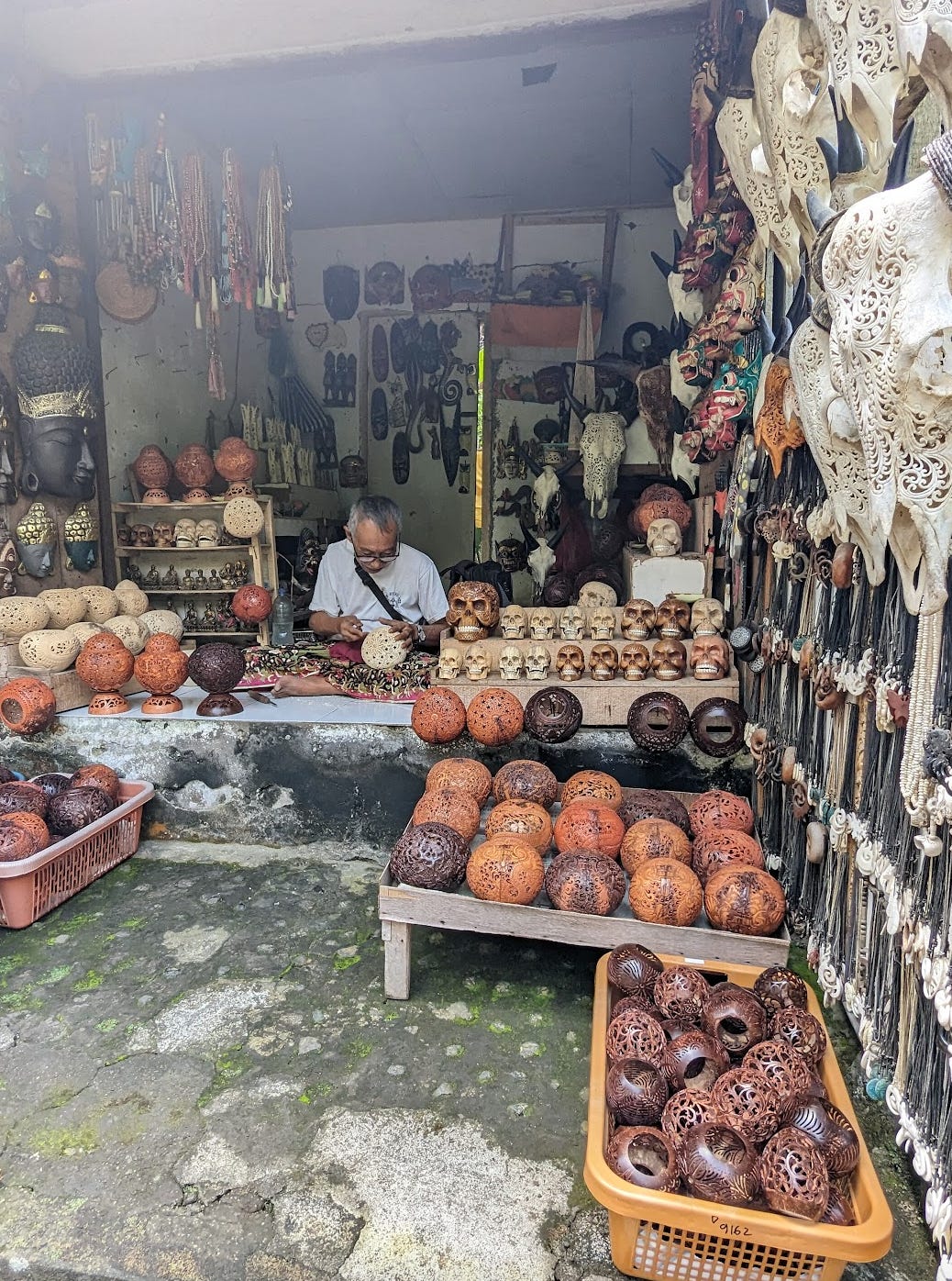 An old man is focused on creating small, tedious, and intricate shapes and designs on a piece of round wood. He is surrounded by his other wood pieces he handmade