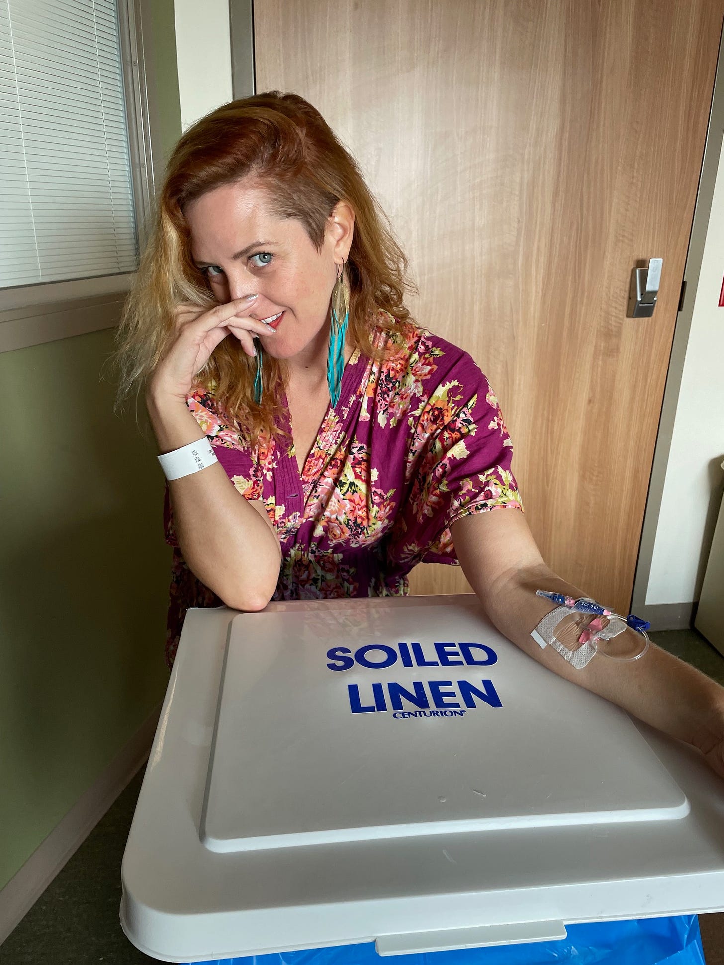 A thin white woman with red hair wears a floral dressing gown and rests her arms atop a container marked "soiled linens" while looking cheekily at the camera.