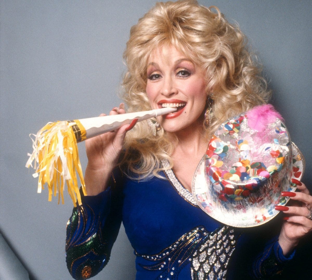 Image of Dolly--a woman smilling, holding a noisemaker to her mouth and a bright derby party hat. She wears a big blonde wig and is smiling