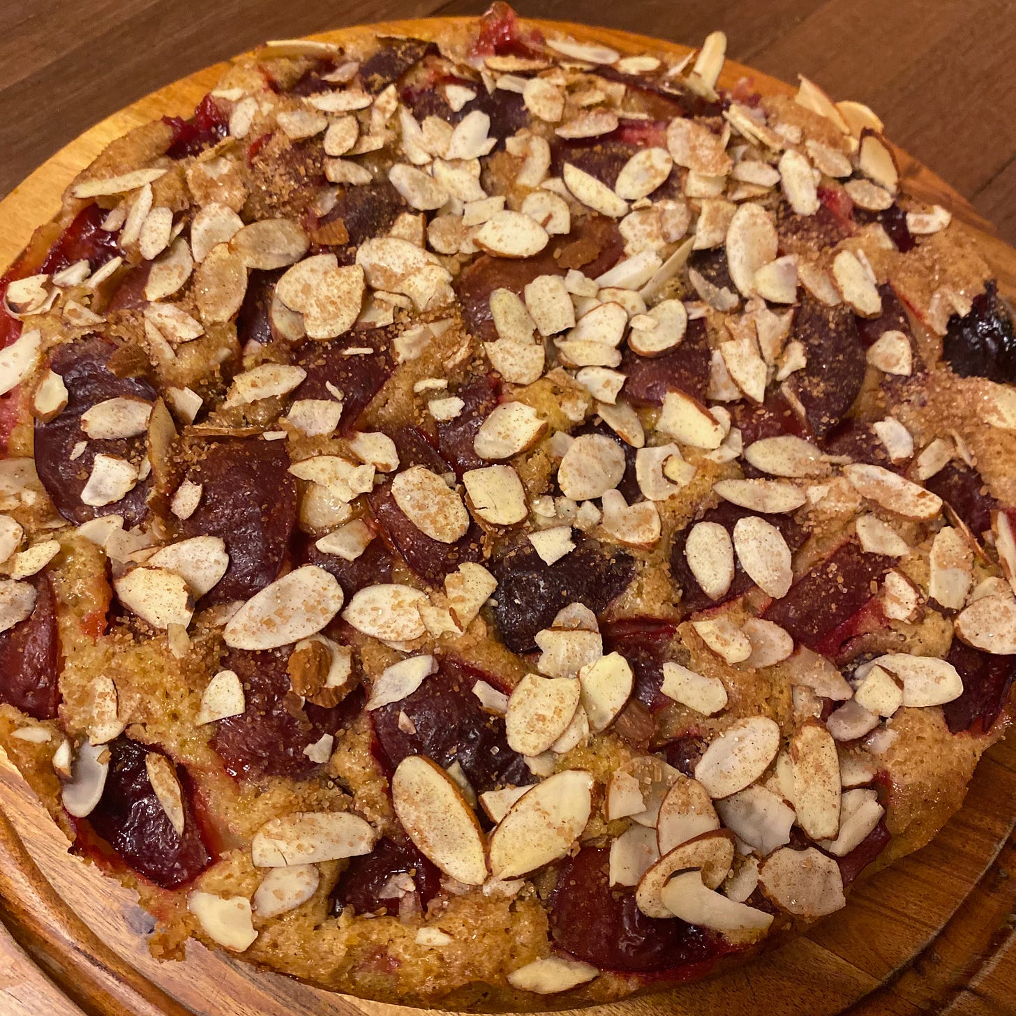 A round cake on a wooden cake board topped with sliced almonds, cinnamon sugar, and deep red plums.