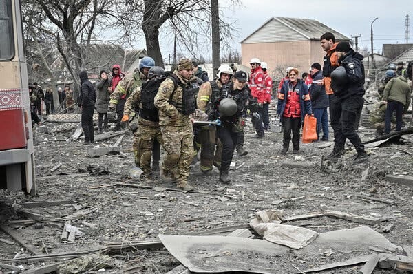 A group of people in military and paramedic uniforms carry a stretcher through a flattened area of rubble.