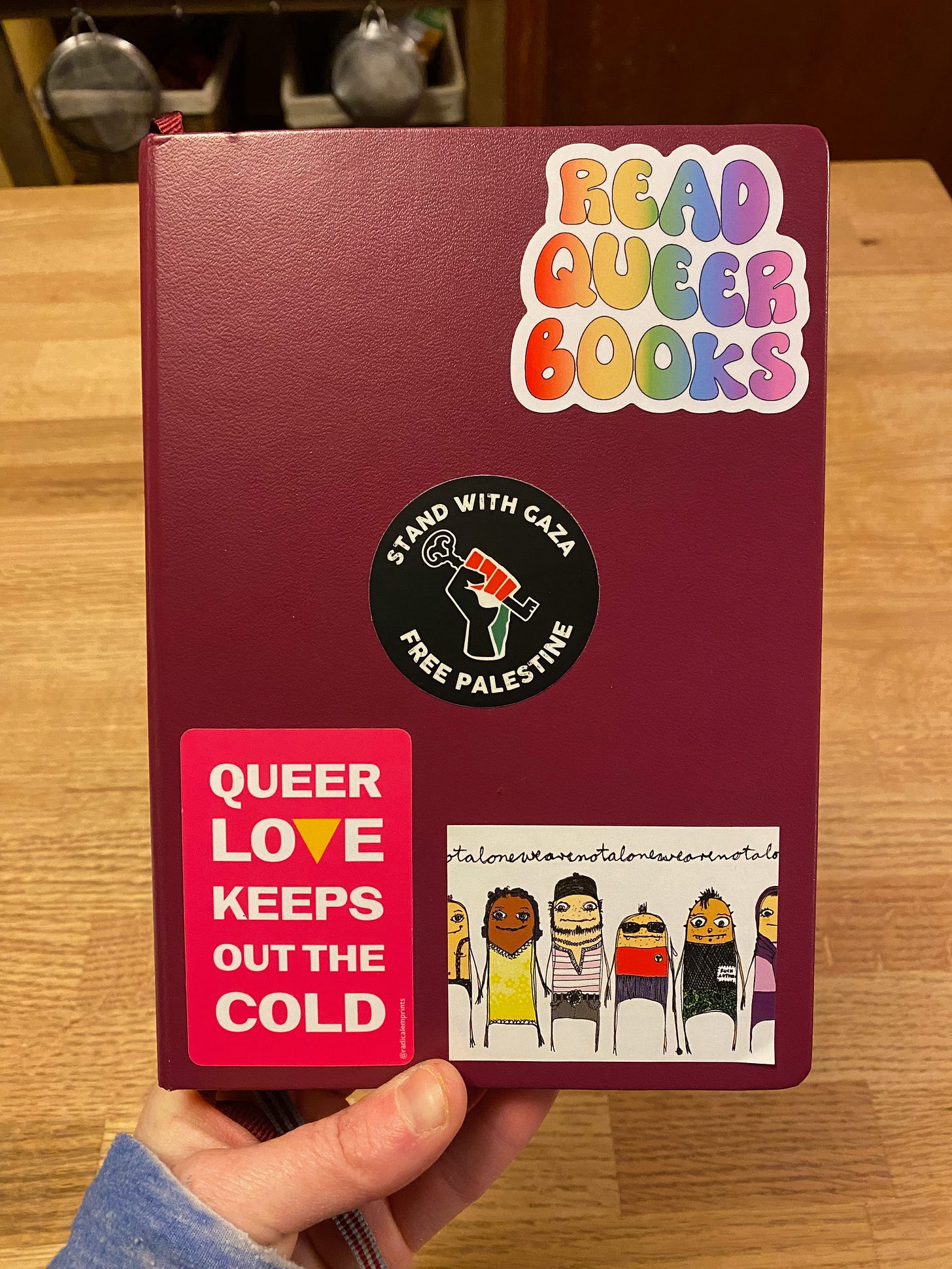 A red planner covered in stickers: Read Queer books in rainbow letters; Stand with Gaza, Free Palestine; Queer Love Keeps Out the Cold in a pink rectangle; and a rectangle of whimsical people wearing colorful clothes above the repeated text: We are not alone.