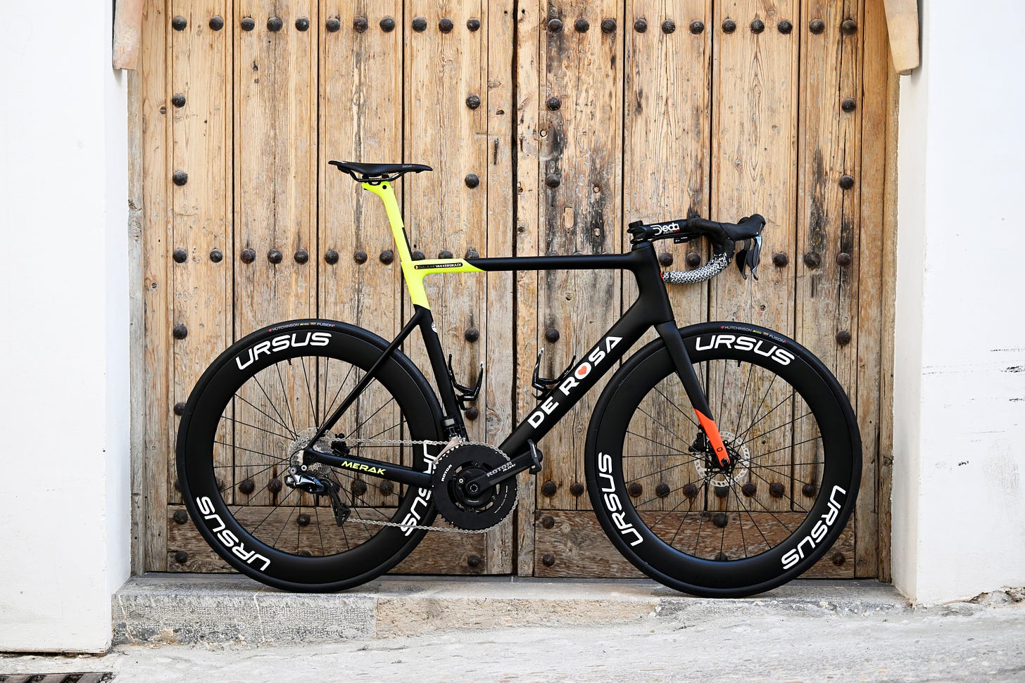 The 2023 De Rosa Merak of Bingoal Wallonie Bruxelles. It is topped off with Ursus 67s, Rotor Aldu crankset, Hutchinson Tyres, Deda Cockpit, Selle Repente saddles, look pedals, swissstop brakes, and Shimano 11-speed groupset, in front of a farm door in the heart of Altea, the heartlands for a cyclist's winter training. 