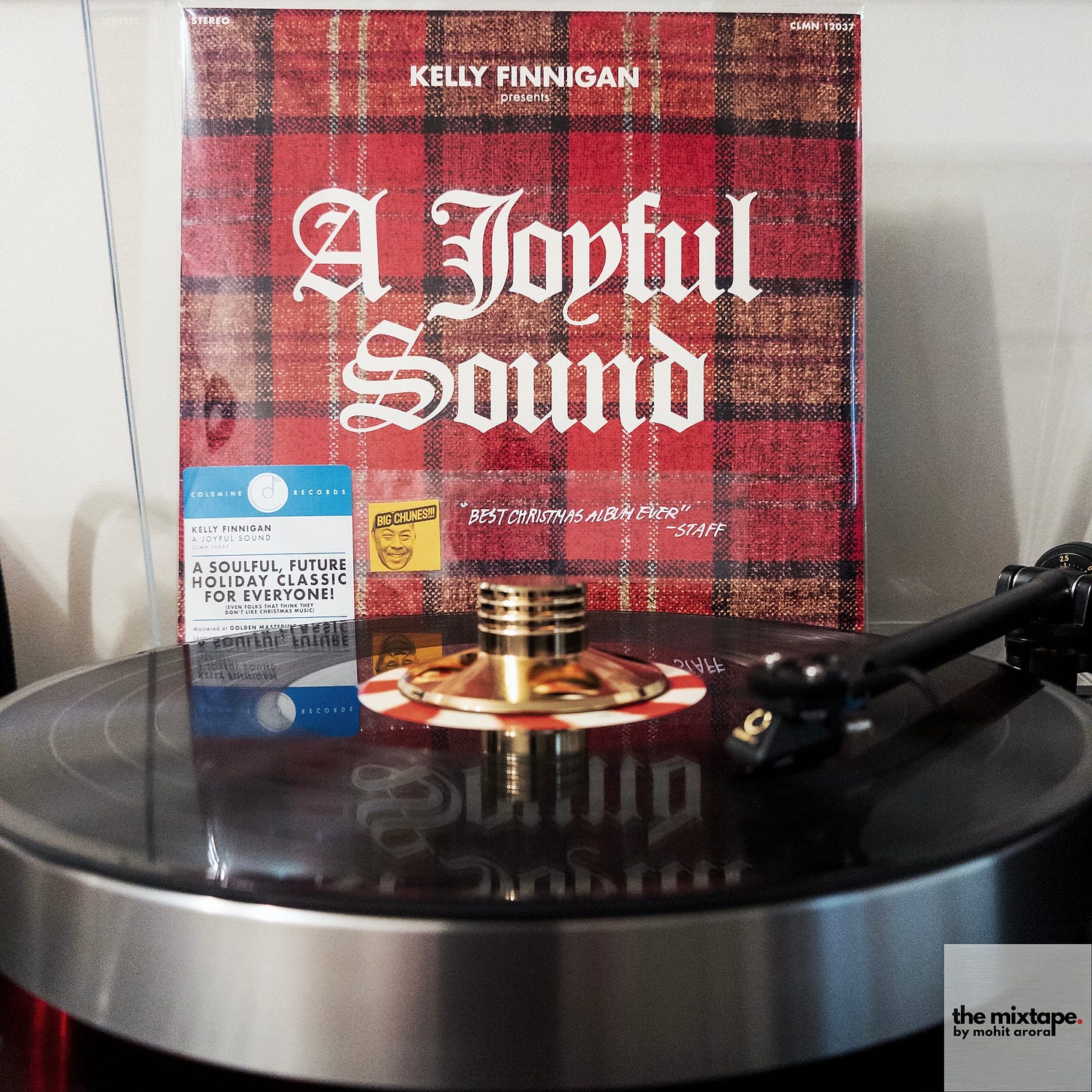 A vinyl record album titled "A Joyful Sound" by Kelly Finnigan on a turntable. The album cover features a red plaid pattern background with gothic-style white lettering. A small sticker on the cover appears to be a staff recommendation. The image shows part of the turntable's metal platter and tonearm, with the black vinyl record visible beneath the clear dust cover.