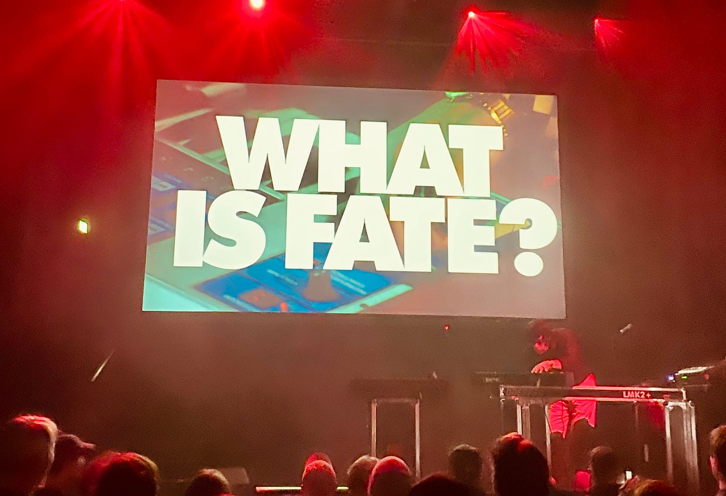 Palco com um músico numa mesa de som e um telão com a frase "What is fate?"