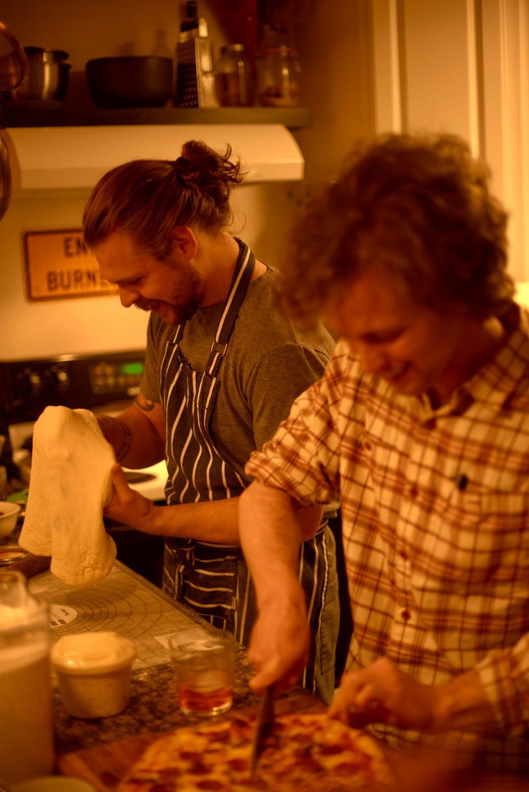 two people standing in a kichen, smiling. one is making pizza dough, and one is cutting into a pizza